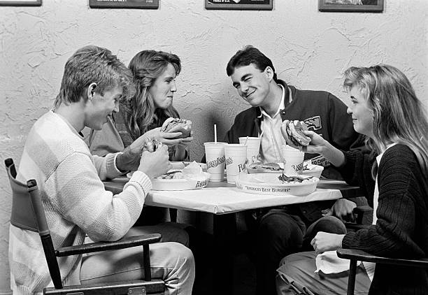 1980s GROUP OF FOUR TEENS SITTING AROUND TABLE AT RESTAURANT CHATTING & EATING HAMBURGERS