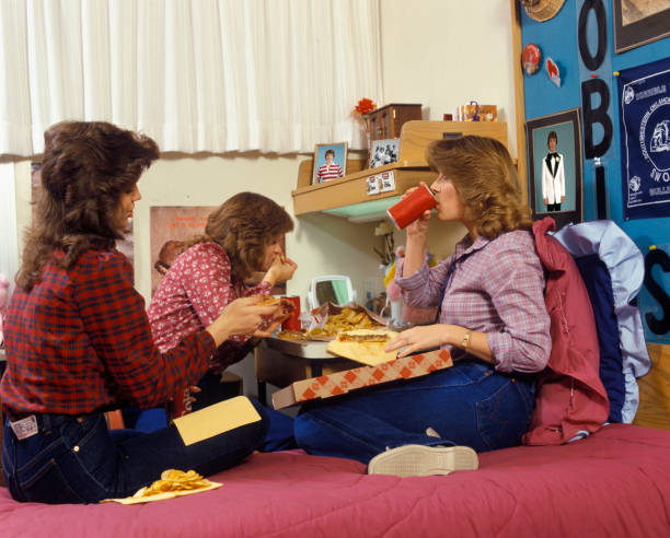 1980s THREE TEENAGE GIRLS EATING PIZZA AND FRENCH FRIES AND DRINKING CANNED SODA IN DORM ROOM