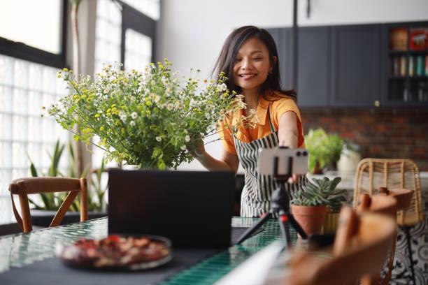 a beautiful asian woman florist is vlogging on her smartphone and laptop, creating an online tutorial on flower bouquet arrangement at home. this scene represents the concept of self-employment, e-learning, online classes, an