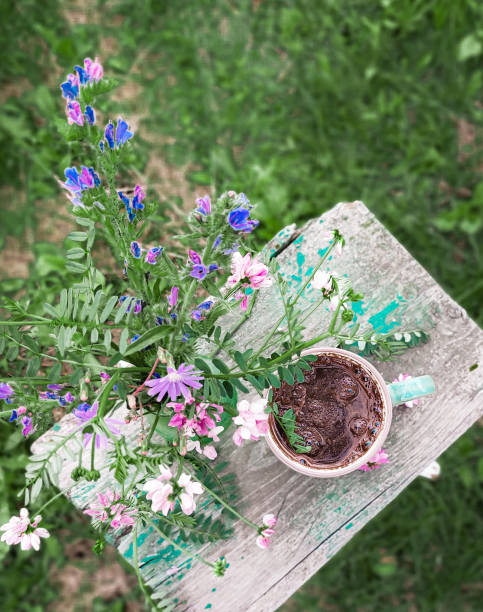 a bouquet of multicolored wildflowers in a small green vase stands on a wooden bench, next to a coffee mug. against the background of a green summer garden - garden decoration stock pictures, royalty-free photos & images