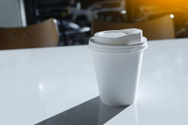 a disposable glass or mug with hot coffee or tea to take away on a white table in a modern stylish youth cafe, cafeteria or restaurant. food and beverage establishment. fast food restaurant. the shadow of the mug. breakfast a