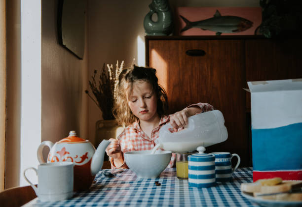 a little girl pours milk on her breakfast cereal in a morning dining table scene - junk food stock pictures, royalty-free photos & images