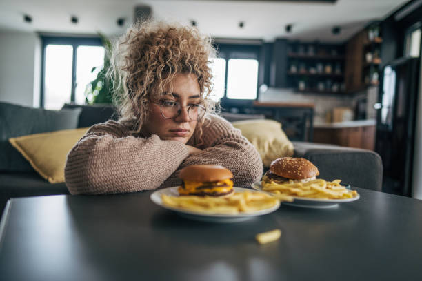 a plus-sized woman with curly hair sitting on the floor, gazing at the two plates of hamburgers and french fries on the table - junk food stock pictures, royalty-free photos & images