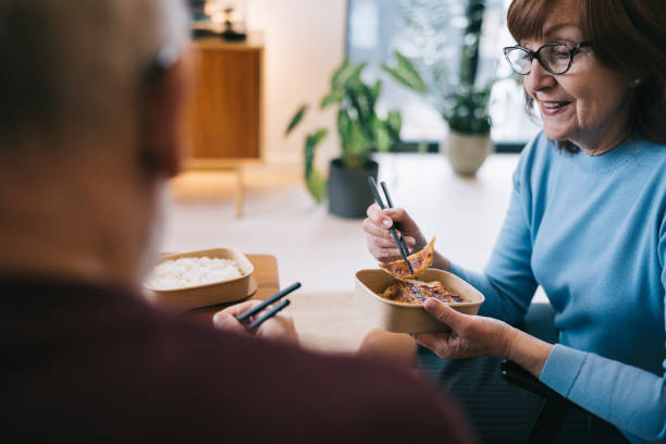 a senior couple shares a cozy moment at home, eating asian takeout food - junk food stock pictures, royalty-free photos & images