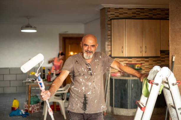 a senior wall painter poses in portrait with tools in hand smiling and looking at camera, full of paint with a roller - home decoration stock pictures, royalty-free photos & images