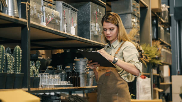 a small business warehouse worker owner wearing apron checking inventory count stocktaking product on shelf warehouse storage room - garden decoration stock pictures, royalty-free photos & images