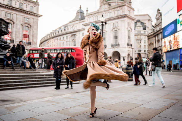 a stylish young woman dressed in 1930s style clothing twirling around by the statue of eros at piccadilly circus - fashion stock-fotos und bilder