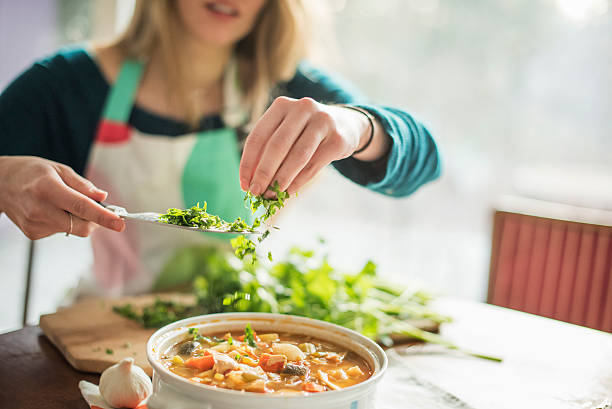 a woman wearing an apron, sitting at a table, sprinkling herbs into a bowl of vegetable stew. - food stock pictures, royalty-free photos & images