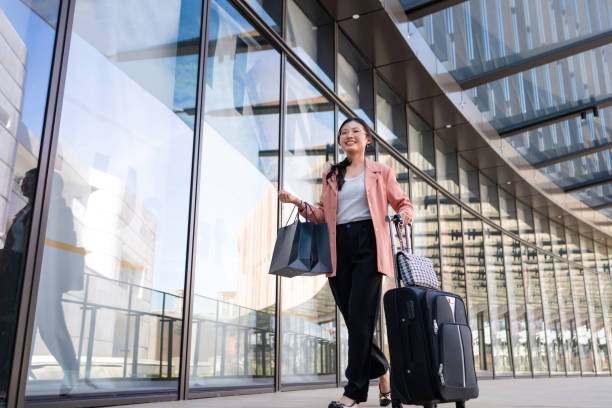 a young beautiful asian business woman with shopping bags in hand while pulling wheeled suitcases at airport terminal - travel stock pictures, royalty-free photos & images