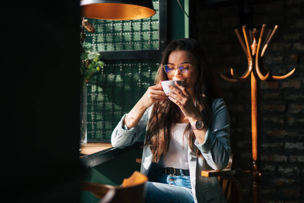 a young beautiful girl is sitting in a cafe by the window and enjoying her coffee. - coffee stock pictures, royalty-free photos & images