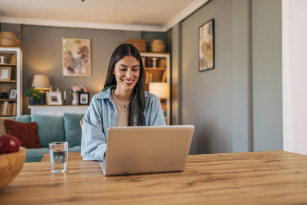 a young cheerful woman sitting at the table in the living room at home and using a laptop. - home decoration photos et images de collection