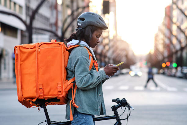 a young delivery woman on her bicycle looking on her smartphone for the route of her delivery - food stock pictures, royalty-free photos & images