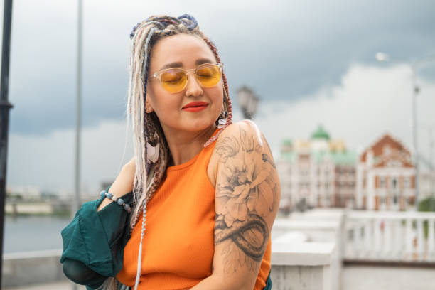 a young happy independent woman with long colored dreadlocks and a tattoo on her shoulder poses on a city street. - fashion stock pictures, royalty-free photos & images
