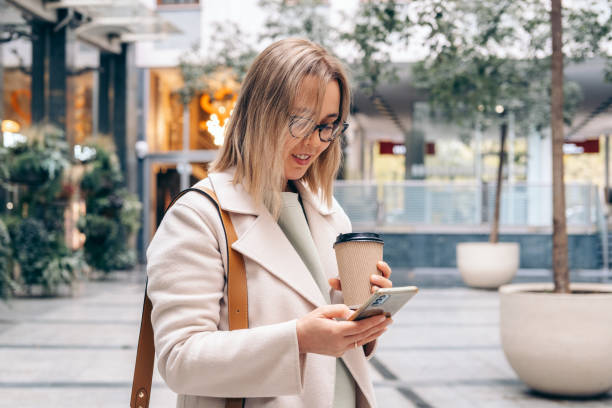 a young woman talking on her smartphone and drinking take-out coffee outdoor - junk food stock pictures, royalty-free photos & images