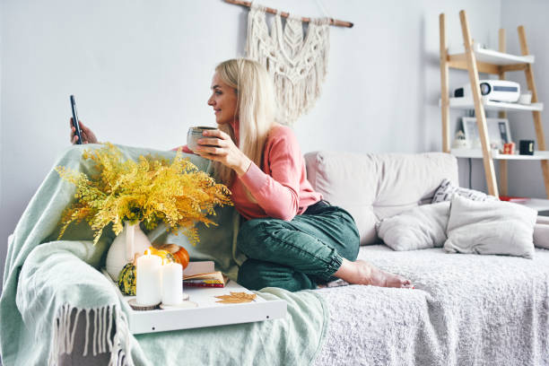 a young woman with blonde hair using mobile phone at home in a cozy atmosphere with autumn decor and pumpkins - home decoration stock pictures, royalty-free photos & images