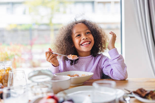 adorable black girl with curly hair smiling while eating breakfast in a bright kitchen - food stock pictures, royalty-free photos & images