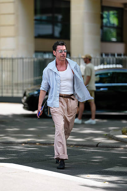 Alex Badia wears silver metallic sunglasses, dark brown beaded necklaces, white tank top, light blue unbuttoned long sleeve shirt, shiny dark brown...