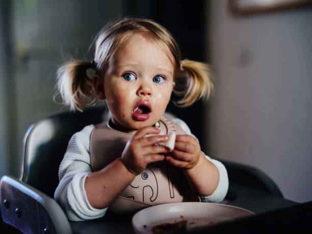 amazed cute toddler girl with pigtails eating at a table at home. - food stock pictures, royalty-free photos & images