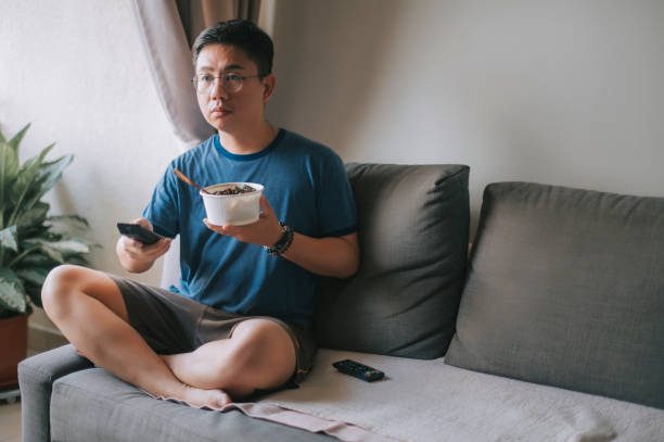 an asian chinese mid adult man having his take out food while pressing tv remote control watching tv on sofa - junk food stock pictures, royalty-free photos & images