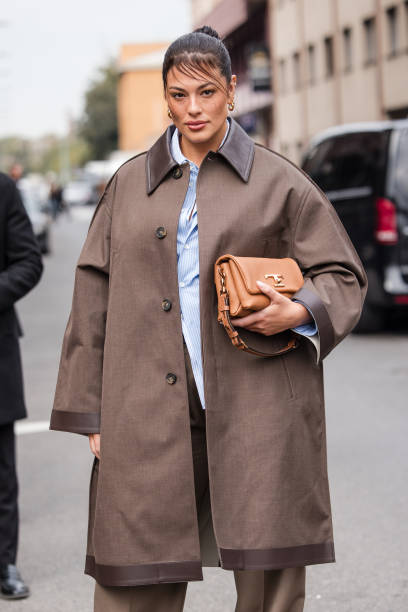 Ashley Graham, wearing a brown trench coat and trousers, a light blue and white striped shirt and and light brown leather bag, is seen outside Tod's...