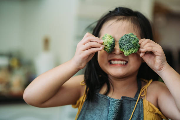asian chinese female child act cute with hand holding broccoli putting in front of her eyes with smiling face at kitchen - food stock pictures, royalty-free photos & images