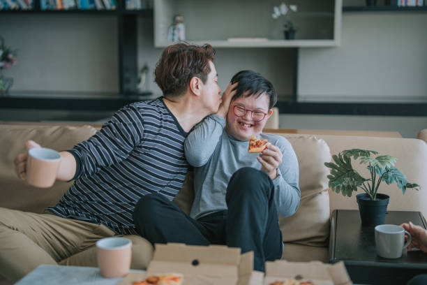 asian chinese man kissing his down syndrome son enjoying pizza fast food in living room sitting on sofa during weekend - junk food stock pictures, royalty-free photos & images