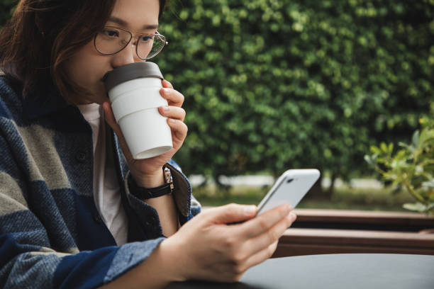 asian woman enjoying coffee using reusable cup while using her phone - junk food stock pictures, royalty-free photos & images