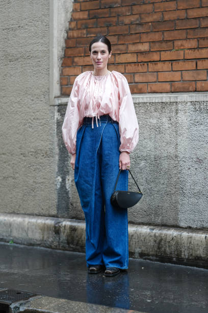 Astrid Boutrot wears silver earrings, light pink long sleeve oversized shirt, navy blue denim jean baggy pants, black Alaïa leather bag, shiny black...