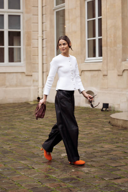 Bambi Northwood-Blyth wears black leather pants, white jacket with buttons, orange high heels and brown Loewe bag outside the Loewe presentation...