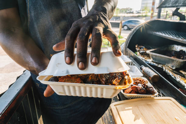 bbq pit chef with a catering company closing a compostabale takeout container full of smoked bbq beans, sausage, chicken thighs & drumsticks, and dirty rice from a food truck or traveling smoker catering company - junk food s