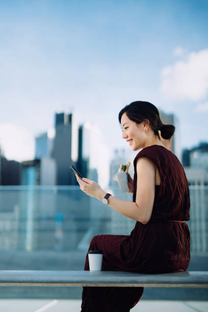 beautiful smiling young asian businesswoman sitting on the bench in office building terrace, against corporate skyscrapers. using smartphone outdoors while eating a healthy sandwich during lunch break. lifestyle and technolog