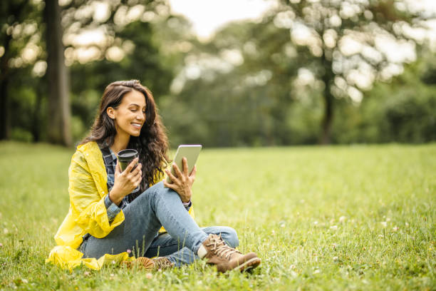 beautiful young lady in yellow raincoat reading a book on her digital tablet, drinking tea and sitting on grass while taking a break in nature - junk food stock pictures, royalty-free photos & images