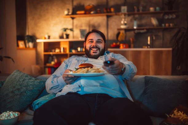übergewichtiger mann essen fast food und vor dem fernseher - junk food stock-fotos und bilder