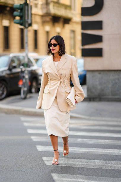 Bettina Looney wears sunglasses, gold earrings, a white shoulder-off blouse, a beige ruffled / epaulets blazer jacket, matching beige leather clutch,...
