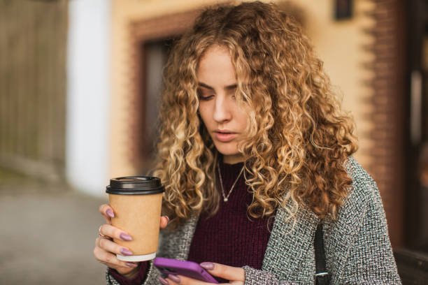 blonde curly girl with paper cup and mobile phone in hand in coat outdoors - junk food stock pictures, royalty-free photos & images
