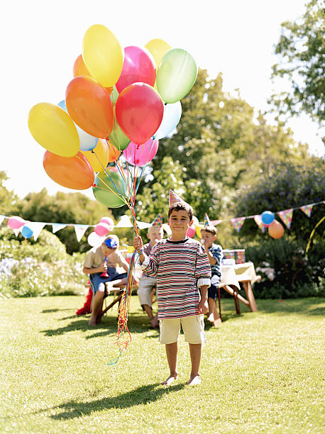 boy (5-7) holding bunch of balloons outdoors, friends in background - garden decoration stock pictures, royalty-free photos & images