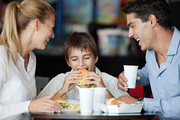 boy eating hamburger in fast food restaurant with his parents - junk food stock pictures, royalty-free photos & images