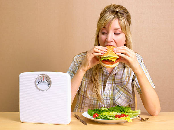 bulimic girl eating burger with bathroom scales - junk food stock pictures, royalty-free photos & images