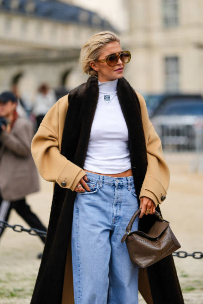Caroline Daur wears dark brown sunglasses, white turtleneck Loewe top, light brown dark brown Loewe coat, light blue denim jean pants, dark green...