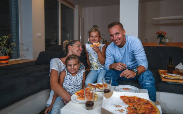 caucasian family sitting on a sofa eating pizza and watching tv - junk food stock pictures, royalty-free photos & images