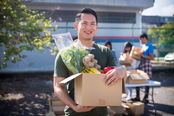 cheerful and smiling asian male volunteer and his colleagues distributing grocery food at community food bank - food stock pictures, royalty-free photos & images