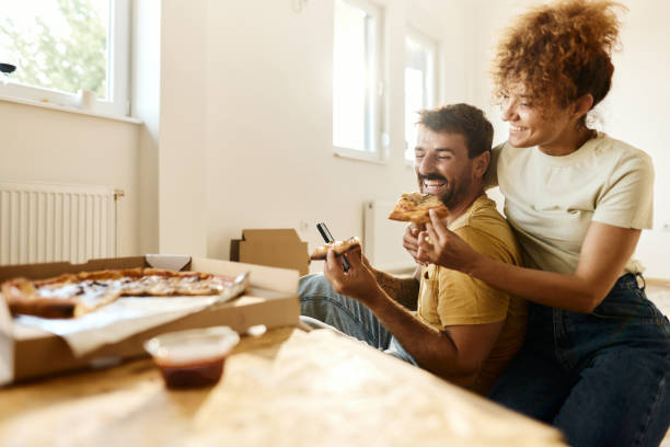 cheerful couple using cell phone and eating pizza on a break from home renovation process. - junk food stock pictures, royalty-free photos & images