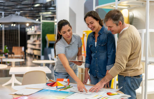 cheerful interior designer holding a tablet and suggesting a color for a home renovation project to customer couple at a furniture store - home decoration stock pictures, royalty-free photos & images