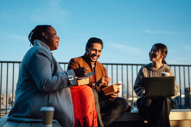 cheerful multi-ethnic business people sitting and having takeaway lunch at rooftop garden at top of office building during lunch break - junk food stock pictures, royalty-free photos & images