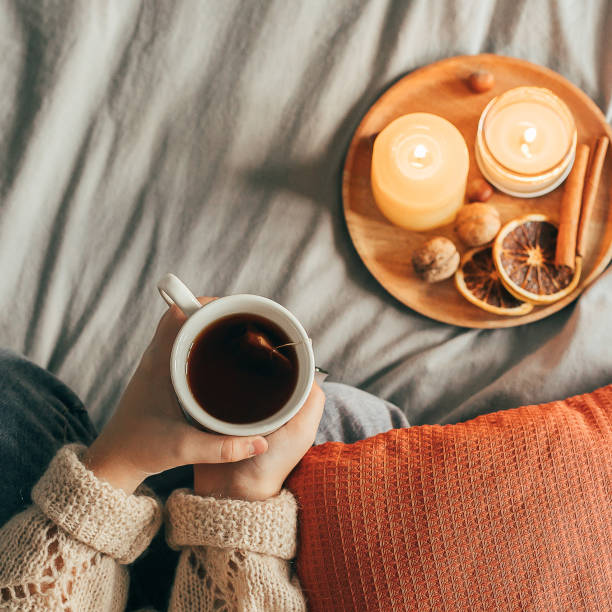 child girl in beige knitted warm sweater drinking hot tea while sitting on bed in bedroom. - home decoration stockfoto's en -beelden