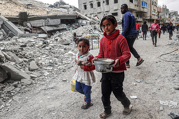 Children walk past the rubble of a collapsed building with a pot of food provided by a charity organisation ahead of the fast-breaking "iftar" meal...