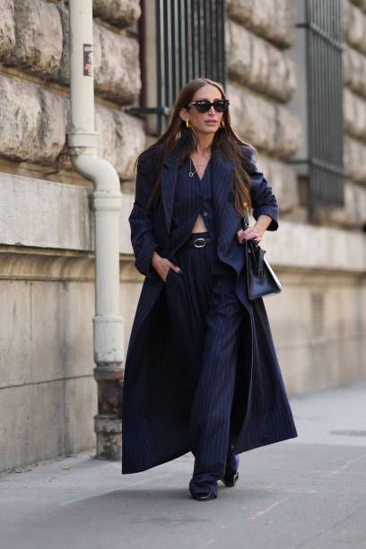 Chloe Harrouche wears sunglasses, a navy blue striped long trench coat, a waistcoat, matching flared suit pants, outside Hermes, during the...