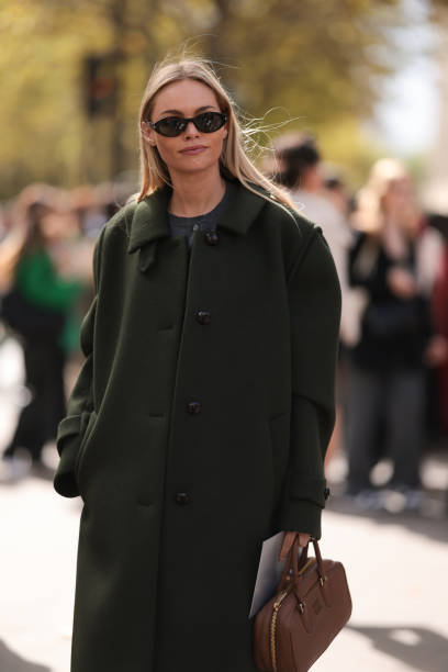Claire Rose Cliteur is seen wearing a long olive green wool coat, black trousers, red patent shoes, black brown oval sunglasses and a brown leather...