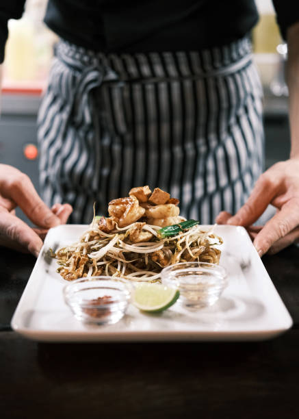 classic thai pad thai noodles with stir-fried shrimp, fresh vegetables, lime, and condiments elegantly presented on a white plate in a traditional restaurant setting - junk food stock pictures, royalty-free photos & images