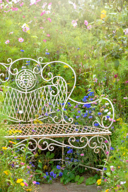 close-up image of a beautiful, vintage style wrought iron garden seat or bench surrounded by summer wild flowers in hazy sunshine - garden decoration stock pictures, royalty-free photos & images